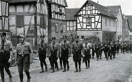 Original-Bildunterschrift: "Propagandamarsch durchs Lumdatal". Die Aufnahme zeigt NS-Einheiten bei einem sog. Propagandamarsch in einem (bisher nicht identifizierten) Ort im Lumdatal. Im Vordergrund mit schwarzen Uniformmützen mit Kokarde SS-Männer, dahinter Angehörige der SA. Die Aufnahme dürfte im Sommer 1931 entstanden sein. [Die mangelnde Bildqualität ist durch die gedruckte Vorlage bedingt.]